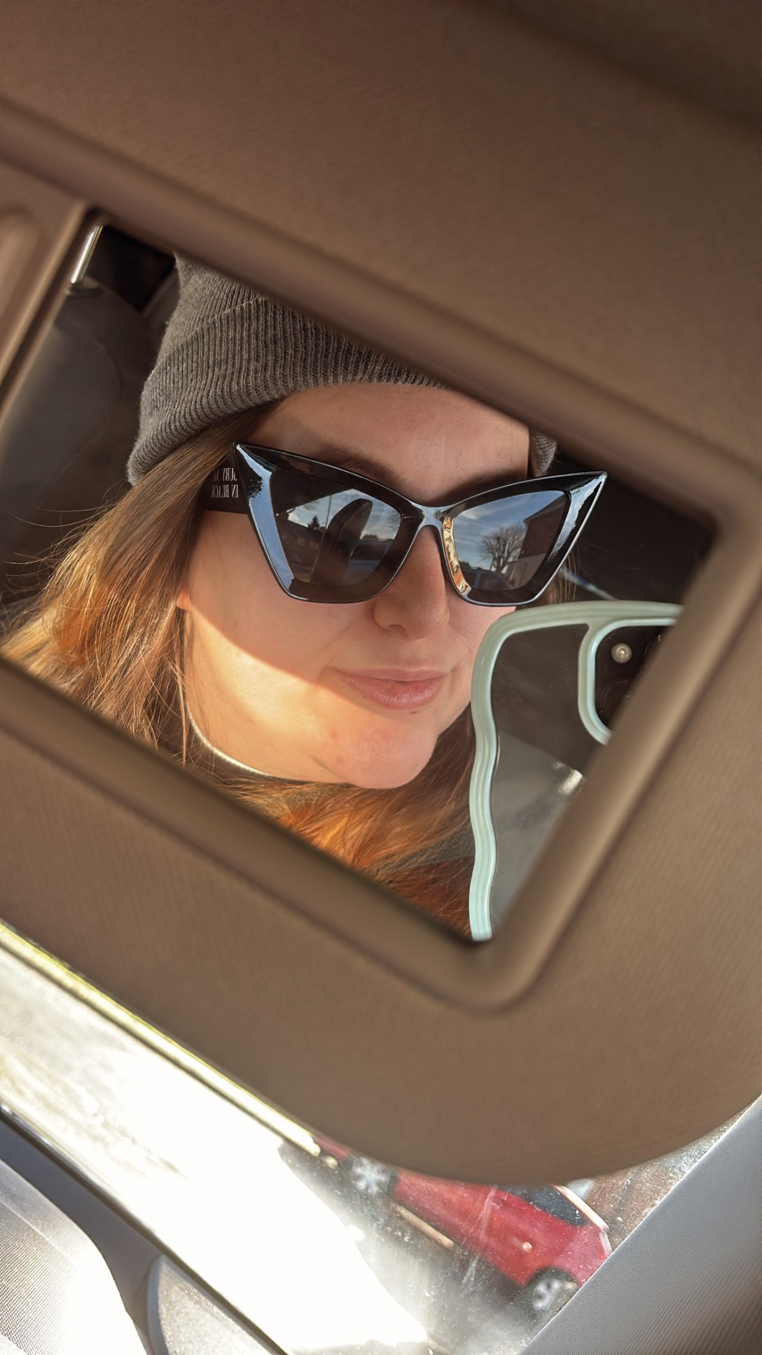 Mirror selfie style picture taken in the car visor mirror. Woman wears black sunglasses, grey beanie and has blonde-brown hair. Her phone peaks in slightly from the right hand side.